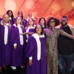 A choir dressed in purple vestments performs on the set of a television show.