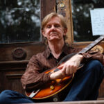 Thomas Jazon holding an electric guitar while sitting down.