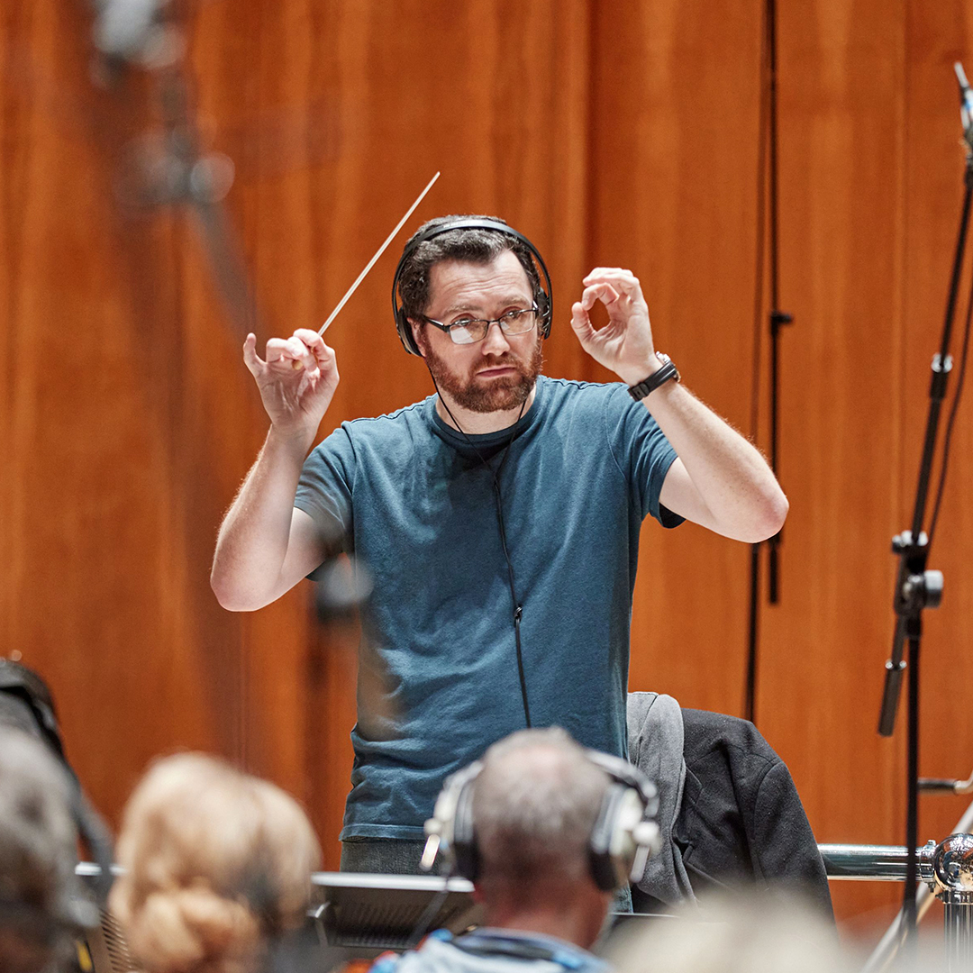 Composer Austin Wintory conducting an orchestra behind a podium on a sound stage.