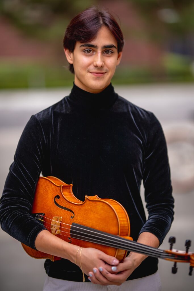 A viola student holding their instrument and smiling on a college campus.