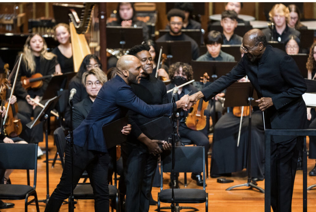 An opera singer shakes the hand of a conductor on the stage of a symphonic concert.