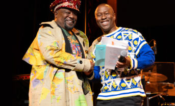 Photo of Dean Jason King smiling and shaking hands with musician George Clinton on the stage of a popular music venue.