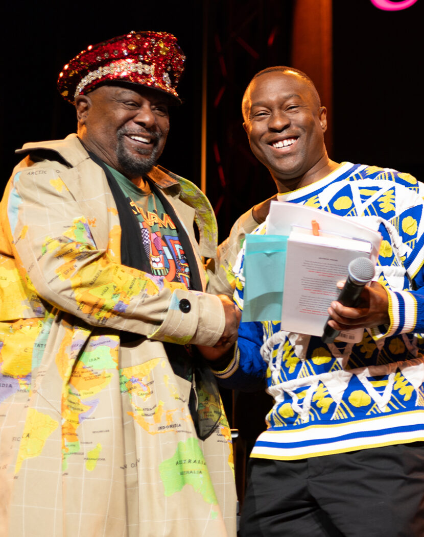 Photo of Dean Jason King smiling and shaking hands with musician George Clinton on the stage of a popular music venue.