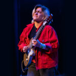 Electric guitarist performs on stage wearing a red jacket.