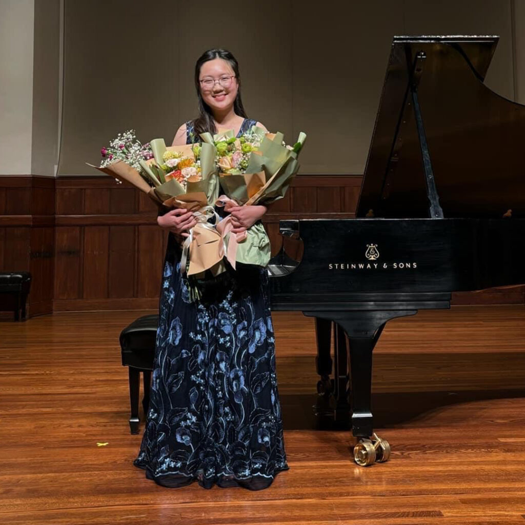 A student musician smiles wearing a blue concert dress with a bouquet of flowers on the stage of a classical theater.