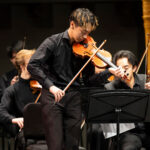 A student violinist plays on a concert stage in dress attire.