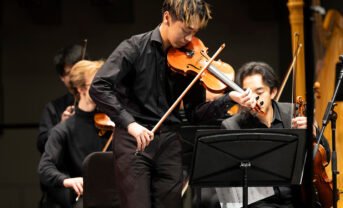 A student violinist plays on a concert stage in dress attire.