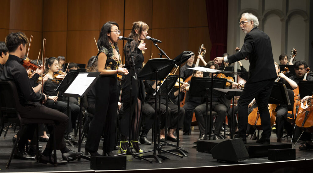A classical music concert is led by a conductor at the podium with two singers.