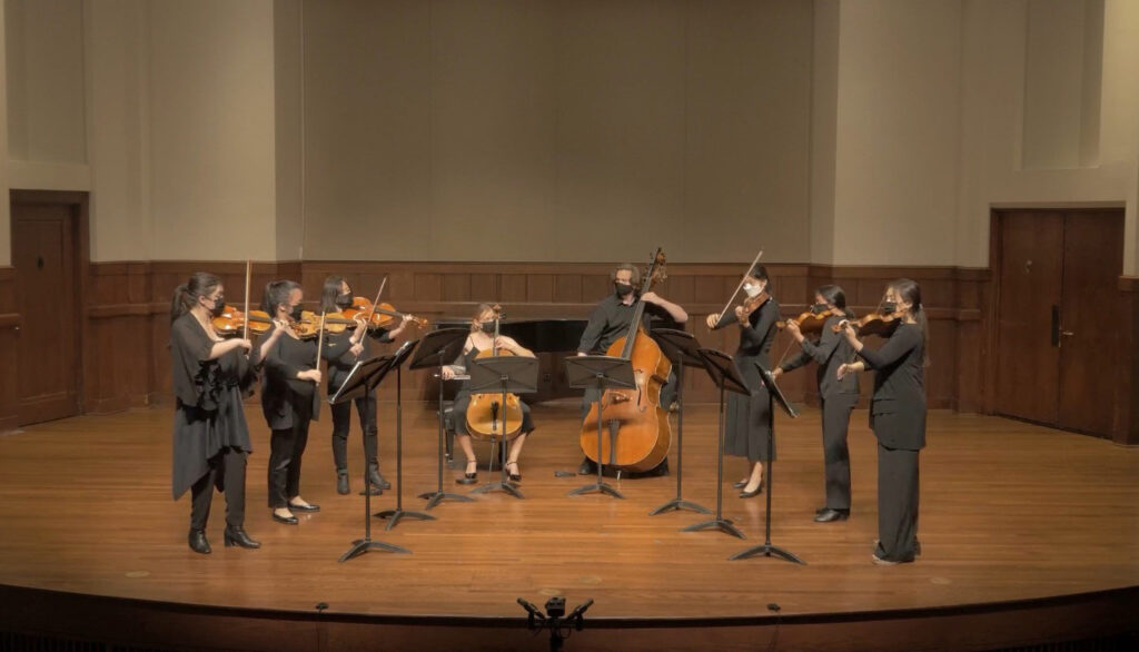 A group of classical strings musicians perform on stage at a recital hall.