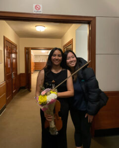 Two classical string musicians smile for the camera outside of a recital hall at a university.