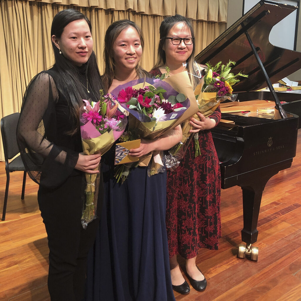 Three classical string musicians smile for the camera holding bouquets of flowers inside a recital hall at a university.