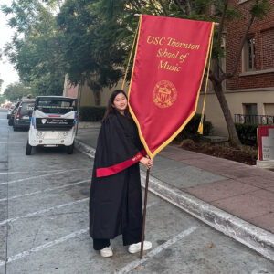 A student in a black graduation robe carries a red ceremonial banner on the campus of a university.