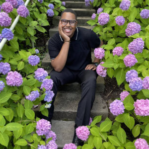 A students smiles at the camera surrounded by purple flowers.