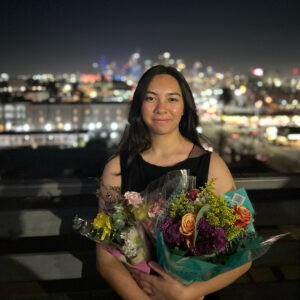 A student wearing a black dress holding a bouquet of flowers smiles at the camera outdoors.