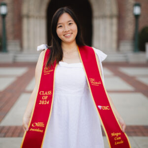 A graduating student in a ceremonial sash smiles at the camera wearing a white dress.