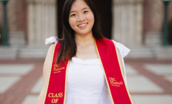 A graduating college student wearing a white dress and a red ceremonial sash smiles at the camera outside a college university.