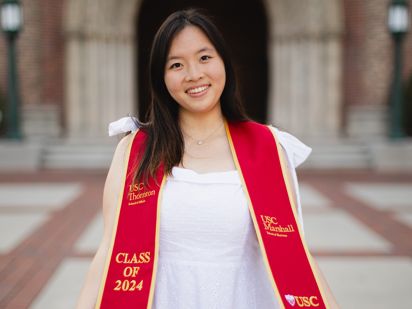 A graduating college student wearing a white dress and a red ceremonial sash smiles at the camera outside a college university. 