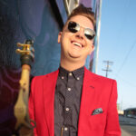 Photo of saxophonist Alex Hahn wearing a red jacket and holding a saxophone outdoors in a city environment.