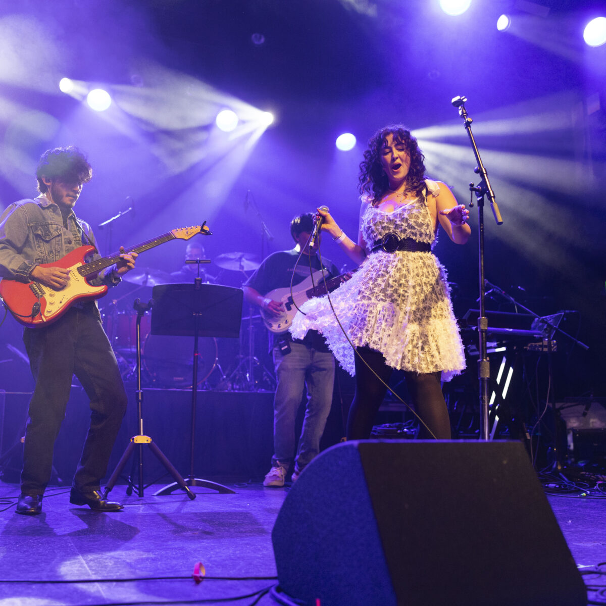 A trio of students sing and play onstage under bright lights.