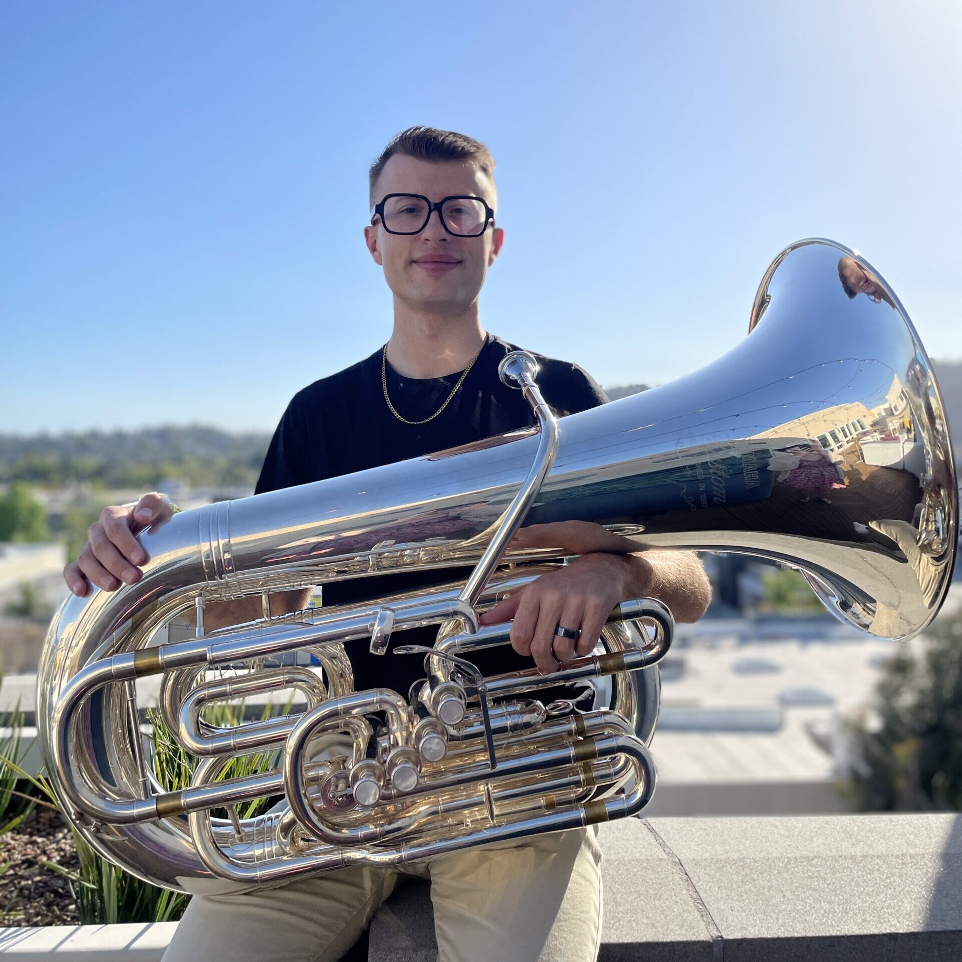 A man with large glasses and a black shirt and gold chain necklace sits outdoors and holds a tuba across his lap