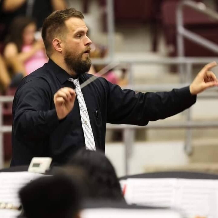 A man in a black shirt and white tie conducts an ensemble onstage