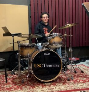 A college-aged student plays at a drum kit.