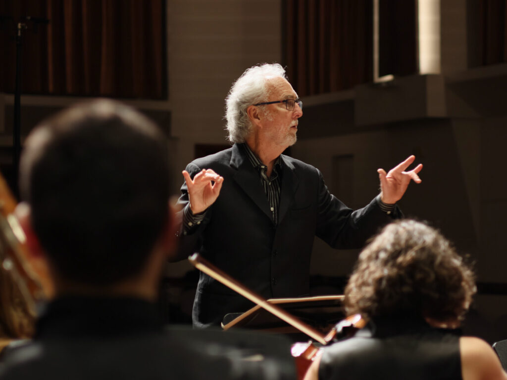A man in a black suit conducts an ensemble with his hands on stage