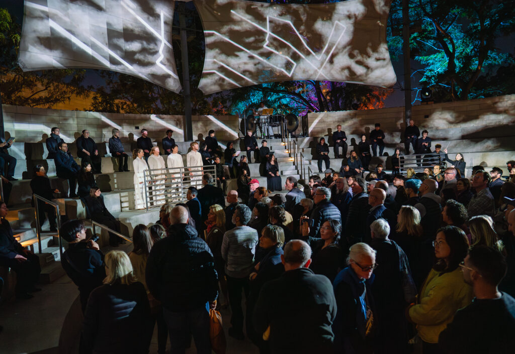 A choir performs outside under a canopy with video projection.