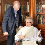 One man sits in a chair, holding a music manuscript, while another stands and looks over the other's shoulder.