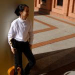 A guitarist poses against a wall, coolly leaning back holding their guitar vertical at their side.