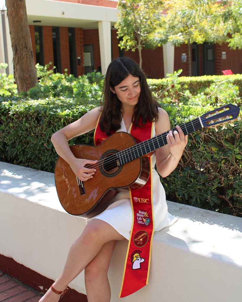Ashley strums an acoustic guitar outdoors in a white dress wearing a decorated Commencement sash.