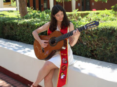 Ashley Melendres plays the guitar, wearing a white dress and decorated Commencement sash.