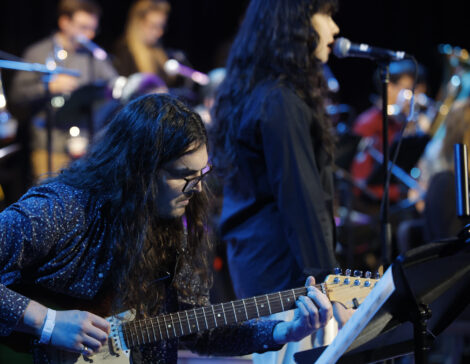 Jazz students perform on electric guitar and vocals surrounded by their bandmates in concert.