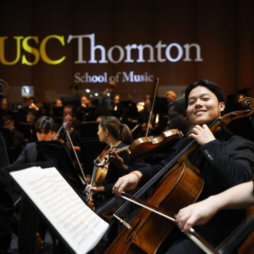 Student cellist smiles at the camera from the orchestra.