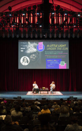 Two panelists sit onstage with a banner behind them in a crowded auditorium.