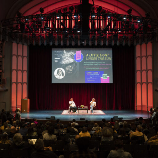 Two panelists sit onstage with a banner behind them in a crowded auditorium.