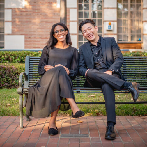 Two students sit on a green park bench, smiling at the camera, wearing concert black.