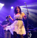 A trio of students sing and play onstage under bright lights.