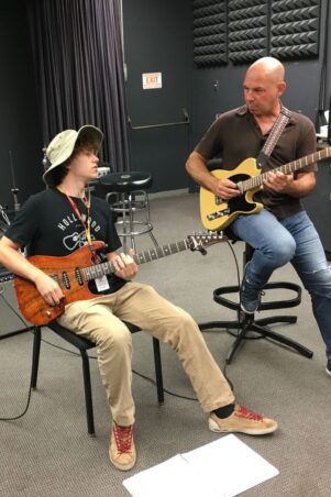 Student and instructor are seated in a music studio, playing guitar.