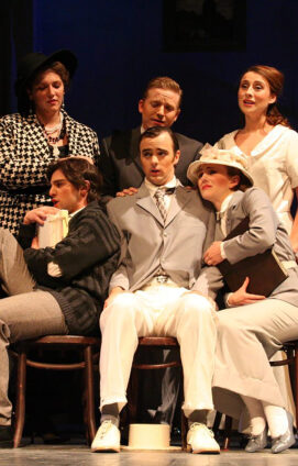 A group of opera cast members sing together onstage wearing vintage attire.