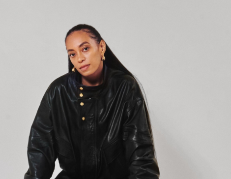 solange seated on a stool, wearing all black against a light grey studio background.
